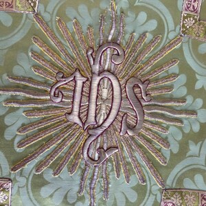 May include: Close-up of a detailed textile with an embroidered design. The central element is the monogram "IHS" in silver thread, surrounded by golden rays. The background is a light green fabric with a subtle floral pattern, and decorative borders.