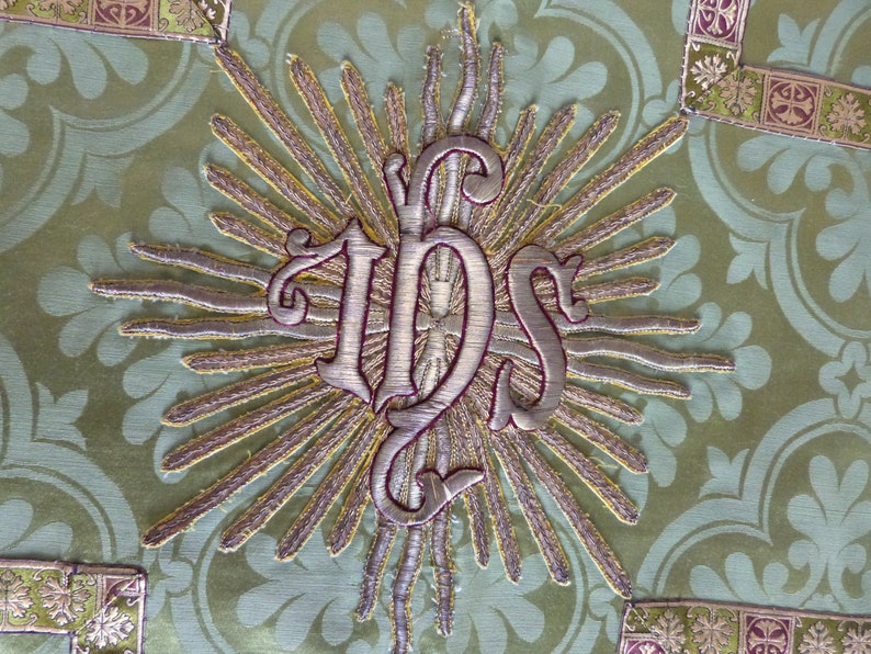 May include: Close-up of a detailed textile with an embroidered design. The central element is the monogram "IHS" in silver thread, surrounded by golden rays. The background is a light green fabric with a subtle floral pattern, and decorative borders.