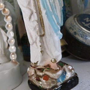 May include: A white figurine of a woman wearing a blue robe with gold accents. The figurine is standing on a black base with painted details.