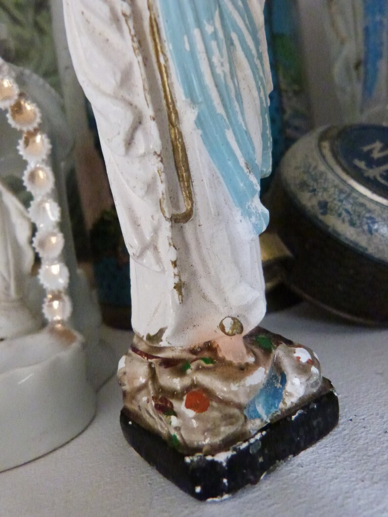 May include: A white figurine of a woman wearing a blue robe with gold accents. The figurine is standing on a black base with painted details.