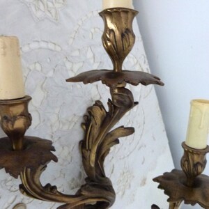 K&ouml;nnte beinhalten: Eine goldfarbene Wandleuchte aus Metall mit drei Kerzenhaltern. Die Leuchte hat ein dekoratives, verziertes Design mit einem floralen Motiv.