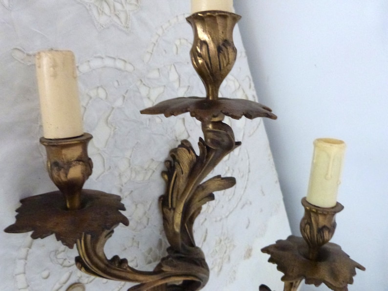 K&ouml;nnte beinhalten: Eine goldfarbene Wandleuchte aus Metall mit drei Kerzenhaltern. Die Leuchte hat ein dekoratives, verziertes Design mit einem floralen Motiv.