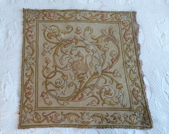 May include: A square, antique tapestry with a detailed design. The tapestry features a central motif of a mythical creature and ornate floral patterns in shades of beige, gold, and pink. The edges have a decorative border.