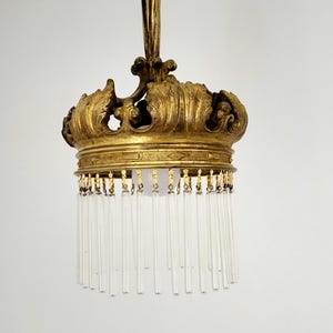 May include: Ornate gold-colored chandelier in a crown shape, featuring a detailed design and hanging clear glass rods. The fixture is suspended from the ceiling, showcasing a vintage aesthetic. The light fixture is a decorative element for interior design.