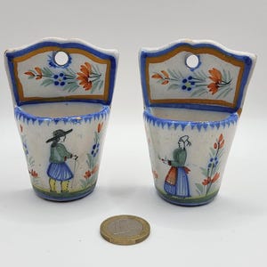 Puede incluir: Dos macetas de pared de cerámica a juego. Cada maceta tiene una base blanca con un borde azul y una parte superior decorativa con un agujero para colgar. Las macetas están adornadas con figuras pintadas a mano y diseños florales en azul, naranja y verde.