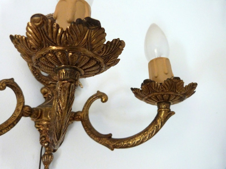 K&ouml;nnte beinhalten: Eine goldfarbene Wandleuchte aus Metall mit zwei Leuchten im Kerzen-Stil. Jede Leuchte hat eine dekorative, verzierte Basis und eine klare Gl&uuml;hbirne.