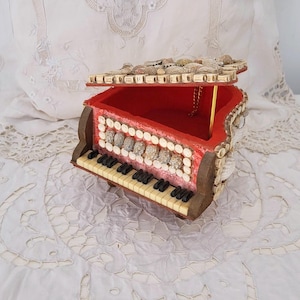 May include: A miniature red piano-shaped music box with a hinged lid. The box features a keyboard with black and white keys, decorative accents, and a gold-colored support rod. The interior is red.