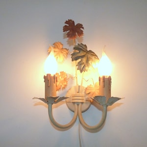 May include: A white metal wall sconce with two candle-shaped lights and a decorative design of leaves in shades of brown, green, and gold.