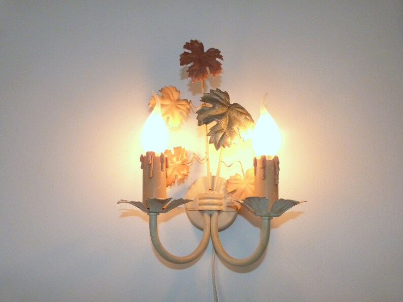 May include: A white metal wall sconce with two candle-shaped lights and a decorative design of leaves in shades of brown, green, and gold.
