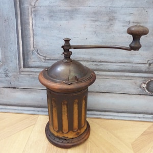 May include: Antique coffee grinder with a wooden base and metal top. The grinder has a hand crank and a metal hopper. The grinder is painted in shades of brown and yellow.