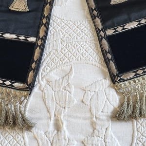 May include: Two black and gold decorative stoles with a rectangular black velvet panel and gold fringe. The stoles have a geometric design with gold and white accents. The stoles are displayed on a white, textured surface.