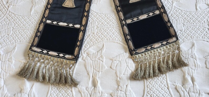 May include: Two black and gold decorative stoles with a rectangular black velvet panel and gold fringe. The stoles have a geometric design with gold and white accents. The stoles are displayed on a white, textured surface.