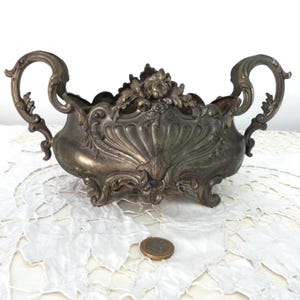 May include: An antique silver-toned decorative bowl with ornate detailing. The bowl features a central floral design and curved handles. The bowl is resting on a white lace tablecloth, with a coin visible in the foreground.
