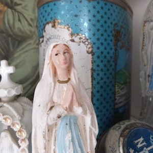 May include: A white porcelain figurine of the Virgin Mary, wearing a blue robe and a white veil. She is holding her hands together in prayer. The figurine is standing on a shelf with other religious items.