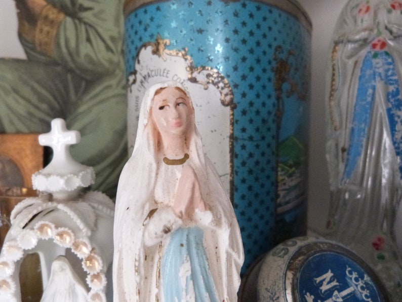 May include: A white porcelain figurine of the Virgin Mary, wearing a blue robe and a white veil. She is holding her hands together in prayer. The figurine is standing on a shelf with other religious items.