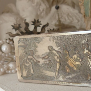 May include: A silver rectangular box with an engraved image of people in a woodland setting. The box has the text "Le Colin-Maillard" and "E. Wiener & Co." on the front.