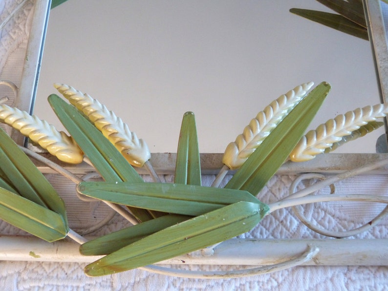 May include: A white metal mirror with a decorative design of green leaves and yellow wheat stalks.
