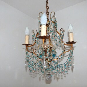 May include: A vintage chandelier with a gold-toned frame, adorned with clear and turquoise-colored crystal beads and teardrop pendants. The chandelier has three candle-style light fixtures with white bulbs, creating a classic and elegant lighting fixture.