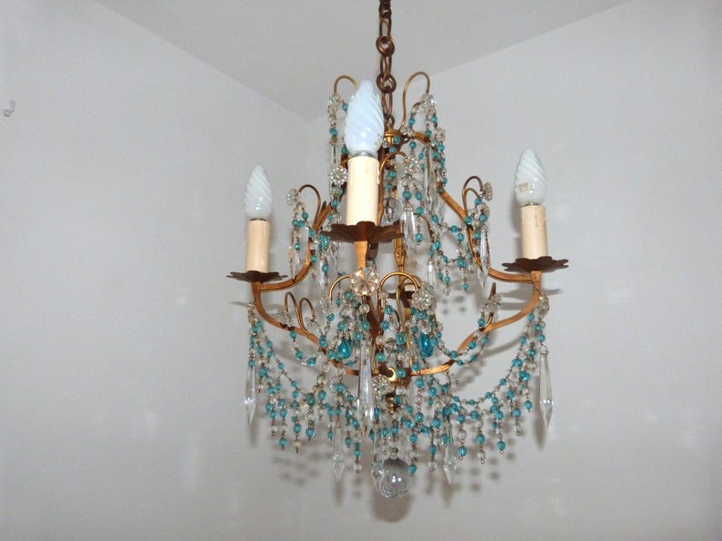 May include: A vintage chandelier with a gold-toned frame, adorned with clear and turquoise-colored crystal beads and teardrop pendants. The chandelier has three candle-style light fixtures with white bulbs, creating a classic and elegant lighting fixture.