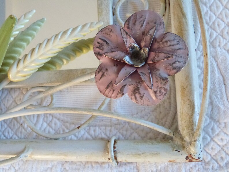 May include: White metal wall mirror with a pink flower and green leaves. The mirror has a decorative, ornate design.