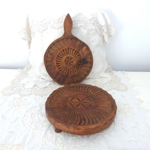 May include: Two intricately carved wooden pieces. One is a round cutting board with a handle, and the other is a small, round, footed plate. Both have a spiral and starburst design. The wood has a warm, brown tone.
