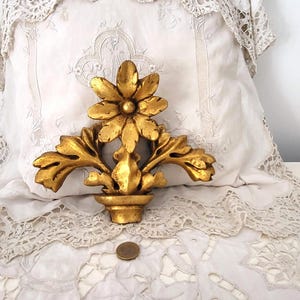 Può includere: Un elemento decorativo floreale dorato e ornato. Il pezzo presenta un fiore centrale con foglie circostanti ed è posto su uno sfondo bianco bordato di pizzo. Una piccola moneta è posta sotto per la scala.