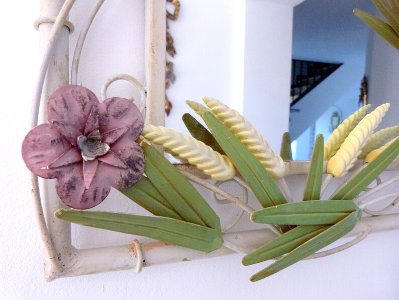 May include: White metal mirror with a floral design. The mirror has a pink flower, green leaves, and yellow wheat stalks.