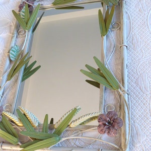 May include: White metal mirror with a decorative floral design. The frame is adorned with green and yellow leaves and a pink flower.