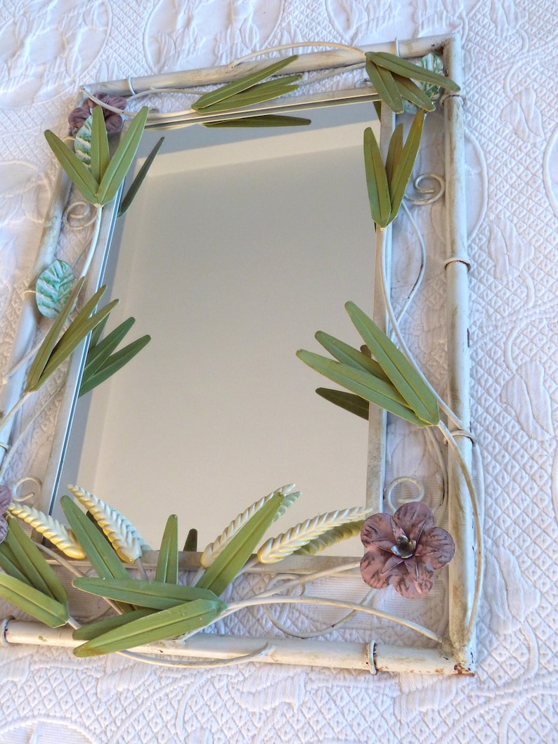 May include: White metal mirror with a decorative floral design. The frame is adorned with green and yellow leaves and a pink flower.
