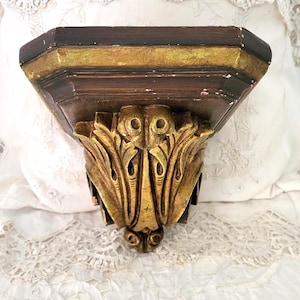 May include: A vintage wall shelf with a dark brown top and a gold-coloured decorative front. The shelf has a detailed, ornate design and is likely made of wood. The gold accents add a touch of elegance to the piece.