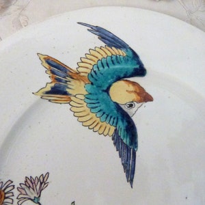 Pode incluir: Um prato de cer&acirc;mica branco com um desenho de p&aacute;ssaro pintado &agrave; m&atilde;o em azul, amarelo e verde. O p&aacute;ssaro est&aacute; em voo e tem uma cauda longa.