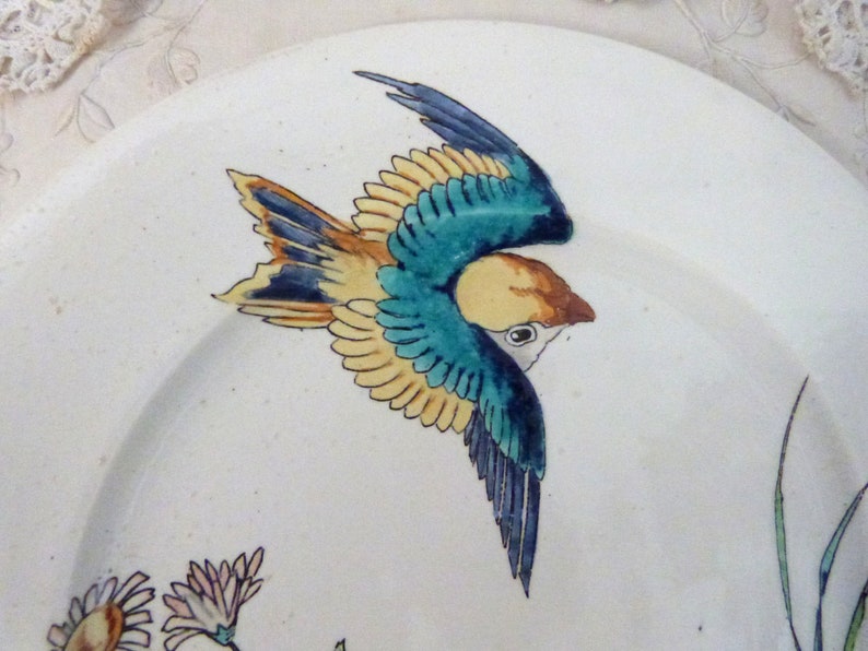 Pode incluir: Um prato de cer&acirc;mica branco com um desenho de p&aacute;ssaro pintado &agrave; m&atilde;o em azul, amarelo e verde. O p&aacute;ssaro est&aacute; em voo e tem uma cauda longa.