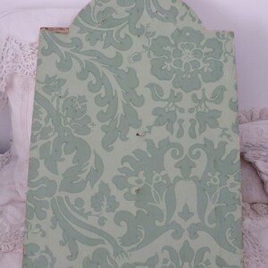 May include: A light green wooden plaque with a decorative floral pattern. The plaque has a rounded top and a metal hook for hanging.
