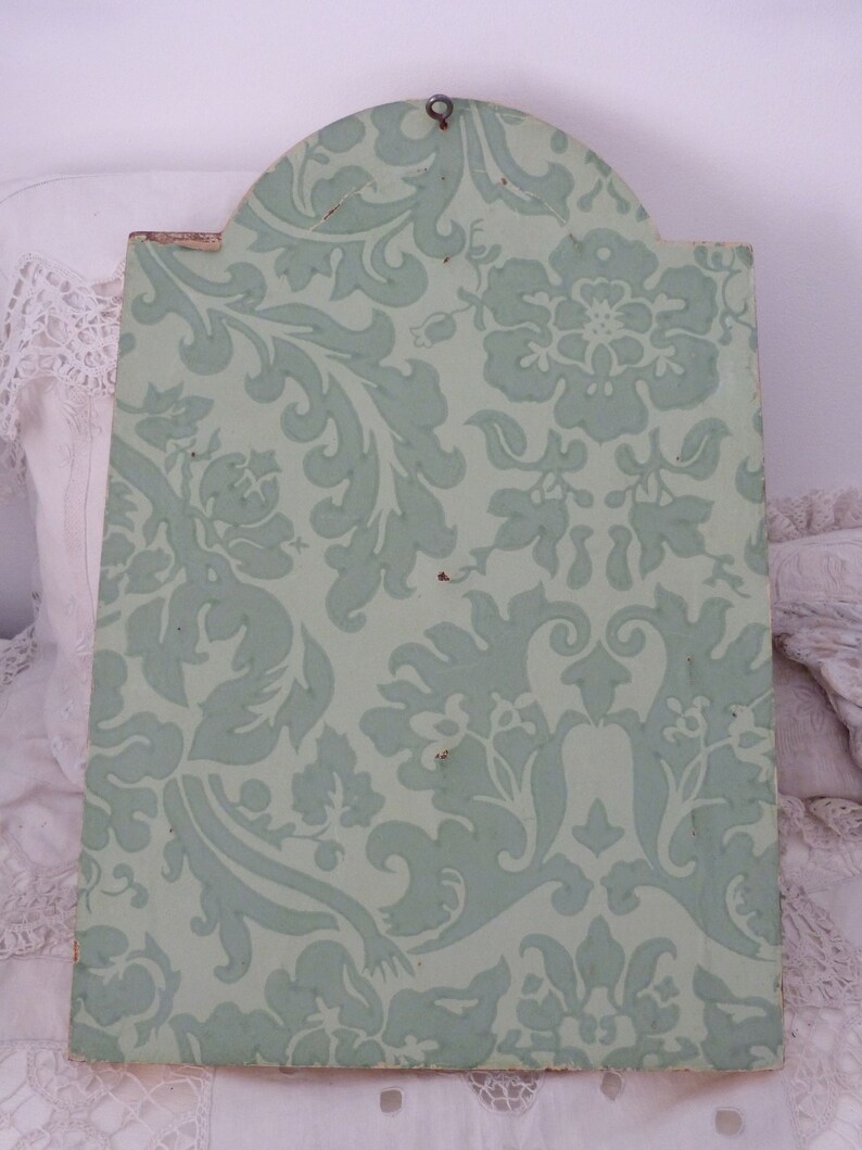 May include: A light green wooden plaque with a decorative floral pattern. The plaque has a rounded top and a metal hook for hanging.