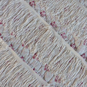 May include: Close-up of white cotton fringe trim. The fringe is thick and fluffy, with a slightly uneven texture.