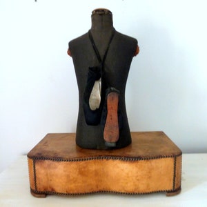 May include: A vintage dress form with a black fabric covering and a wooden base. The dress form is on a brown leather platform with decorative stitching.