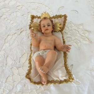 May include: A vintage figurine of the baby Jesus, lying on a white satin pillow with a gold trim. The baby has light skin, brown hair, and is wearing a white diaper. A gold halo is behind the baby's head.