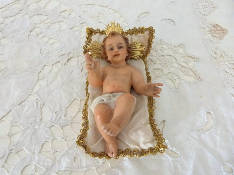 May include: A vintage figurine of the baby Jesus, lying on a white satin pillow with a gold trim. The baby has light skin, brown hair, and is wearing a white diaper. A gold halo is behind the baby's head.