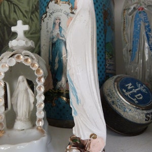 May include: A white porcelain figurine of the Virgin Mary, wearing a blue and white robe, standing on a black base. The figurine is in front of a blue and white tin with stars and the text "Je Suis L'Immaculee Conception".