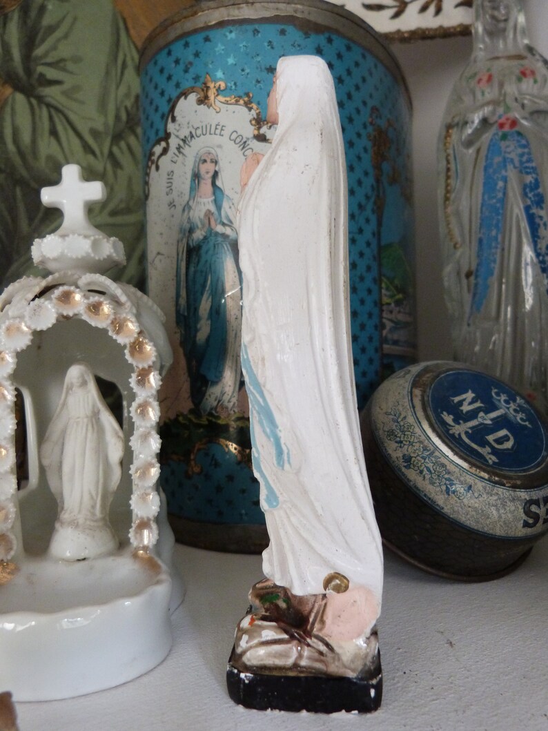 May include: A white porcelain figurine of the Virgin Mary, wearing a blue and white robe, standing on a black base. The figurine is in front of a blue and white tin with stars and the text "Je Suis L'Immaculee Conception".