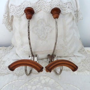 May include: Two wall-mounted coat hooks featuring curved, light brown wooden arms and rounded wooden finials at the top. The vertical supports are made of weathered, tarnished silver-toned metal with visible signs of age and oxidation.