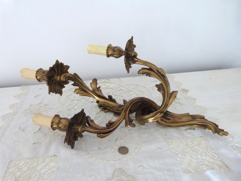 K&ouml;nnte beinhalten: Eine goldfarbene Wandleuchte aus Metall mit zwei Kerzenleuchten. Die Leuchte hat ein dekoratives, verziertes Design mit geschwungenen, blattartigen Details.