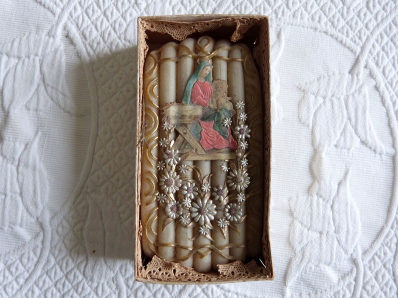 May include: A set of white candles in a box with a religious image of Mary and baby Jesus. The candles are decorated with gold and silver accents and white flowers.
