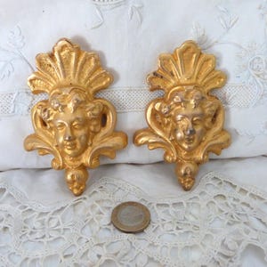 May include: Two gold-toned metal wall decor pieces featuring a stylized face with a shell-like design on top. The pieces are ornate and have a vintage aesthetic.