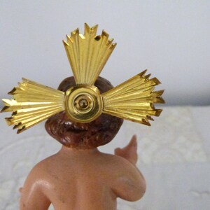 May include: A small figurine of a baby with brown hair and skin, wearing a gold halo. The halo is a cross-shaped design with radiating beams. The figurine is a religious item, likely for a nativity scene or similar display.