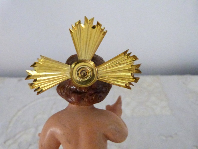 May include: A small figurine of a baby with brown hair and skin, wearing a gold halo. The halo is a cross-shaped design with radiating beams. The figurine is a religious item, likely for a nativity scene or similar display.