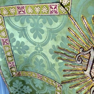 May include: Close-up of a green textile with a floral pattern. The fabric is adorned with decorative trim featuring a repeating design of leaves and geometric shapes in green, purple, and white. A gold embroidered sunburst design is visible.