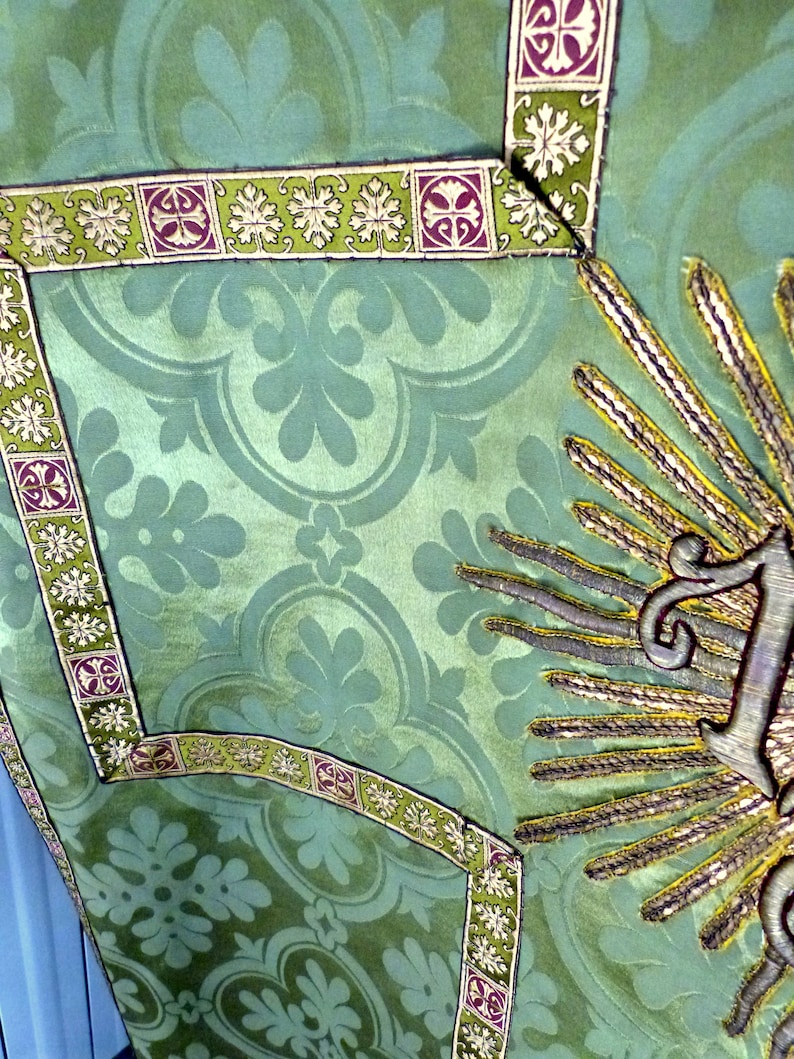 May include: Close-up of a green textile with a floral pattern. The fabric is adorned with decorative trim featuring a repeating design of leaves and geometric shapes in green, purple, and white. A gold embroidered sunburst design is visible.