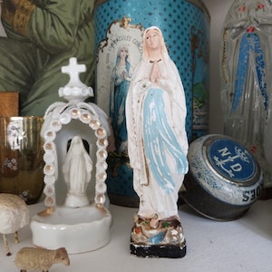 May include: A collection of religious figurines and decorative items, including a white porcelain statue of the Virgin Mary, a blue and white tin box with the words "S.S. IMMACULEE CONCEPTION" and "N.D. DES SEIGES", and a clear glass figurine of the Virgin Mary.
