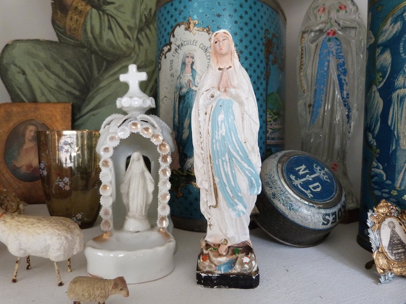 May include: A collection of religious figurines and decorative items, including a white porcelain statue of the Virgin Mary, a blue and white tin box with the words "S.S. IMMACULEE CONCEPTION" and "N.D. DES SEIGES", and a clear glass figurine of the Virgin Mary.